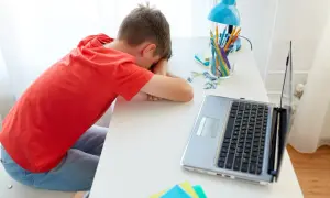 Chłopiec w czerwonej koszulce śpi na biurku obok laptopa i przyborów szkolnych.