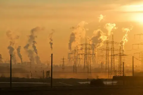 Dymiące kominy fabryk i linie energetyczne na tle zachodzącego słońca symbolizują antropogeniczne zmiany klimatu.