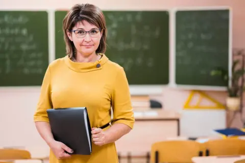 Nauczycielka w okularach, w żółtej sukience, trzyma teczkę. To ona może prowadzić zajęcia rewalidacyjne.