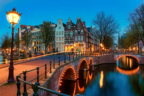 Most Amsterdam canals are a punkt wyjściowy do pomiarów wysokości na lądzie. Widok na kanał z mostem oświetlonym lampkami i kamienicami w tle.