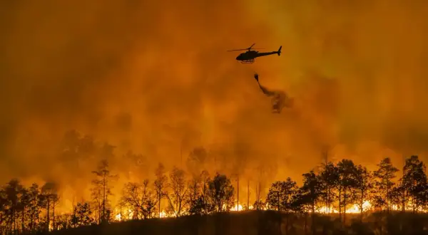 Helikopter zrzuca wodę na płonący las. Intensywny pomarańczowy dym i ogień to przerażające skutki globalnego ocieplenia.