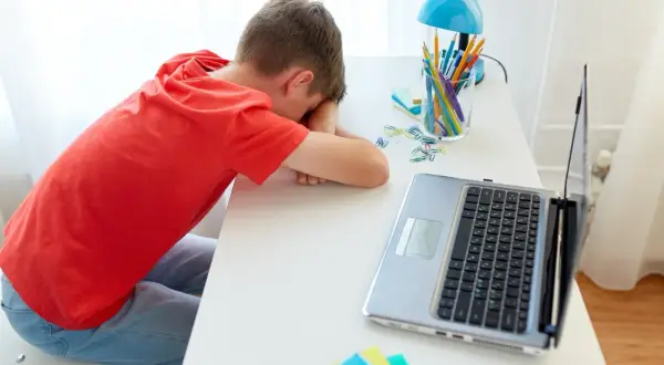 Chłopiec w czerwonej koszulce śpi na biurku obok laptopa i przyborów szkolnych.