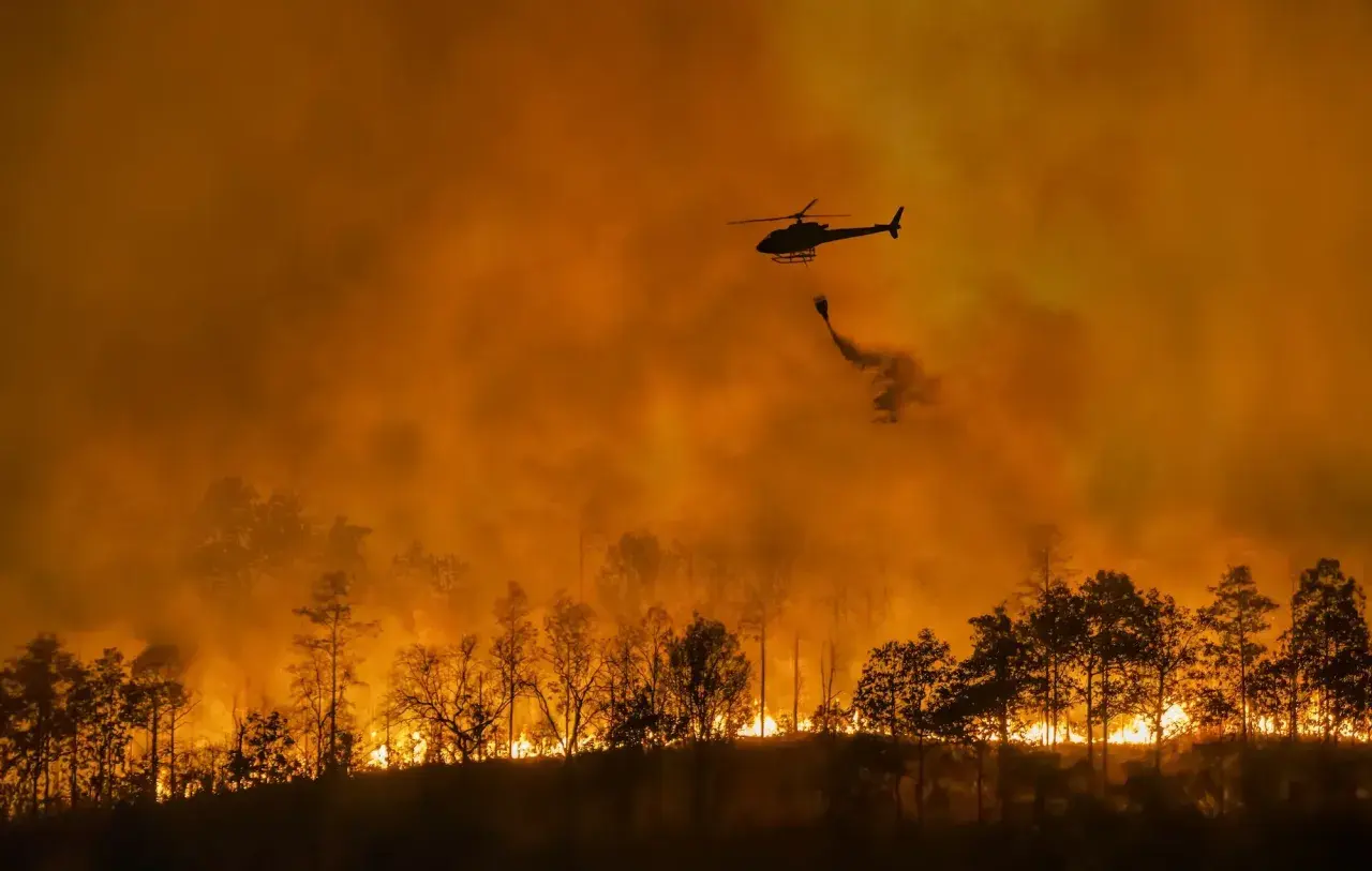 Helikopter zrzuca wodę na płonący las. Intensywny pomarańczowy dym i ogień to przerażające skutki globalnego ocieplenia.