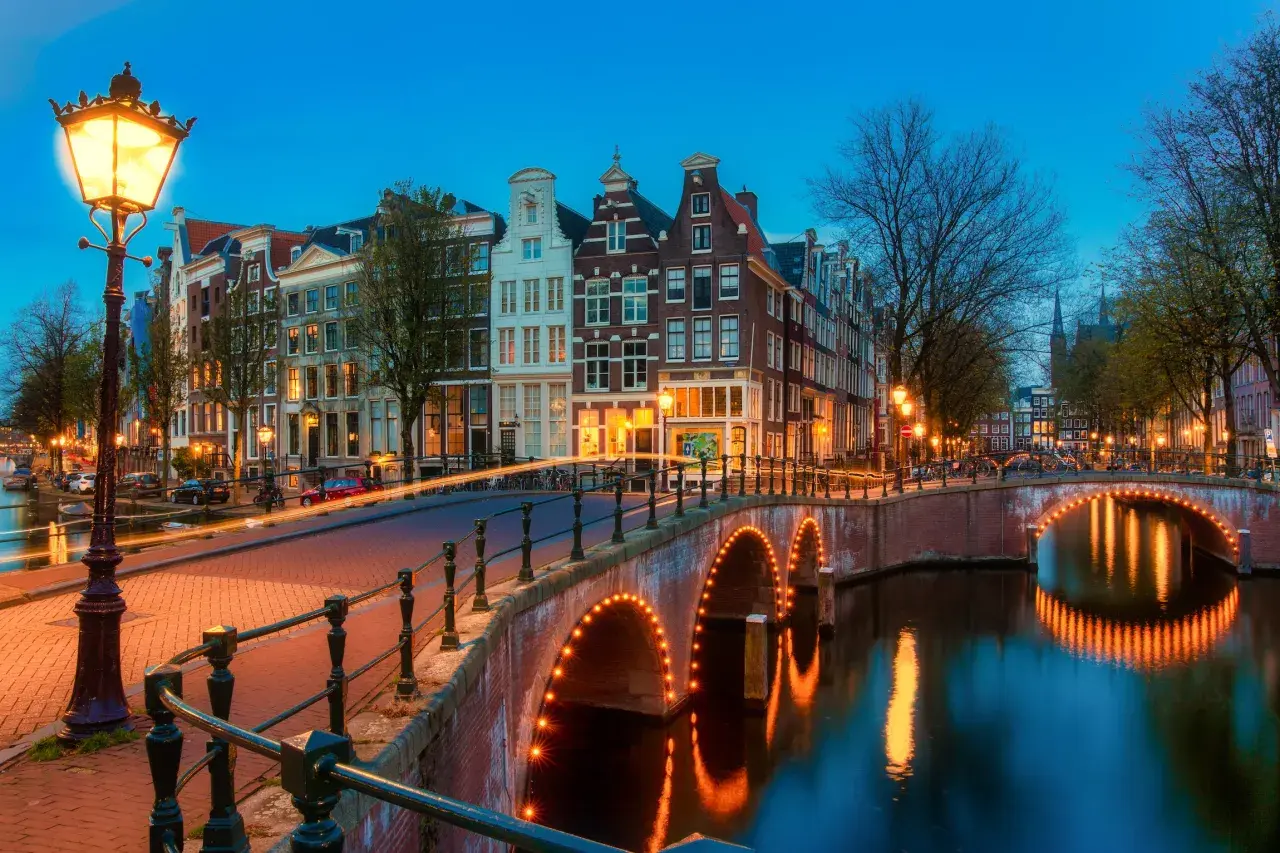 Most Amsterdam canals are a punkt wyjściowy do pomiarów wysokości na lądzie. Widok na kanał z mostem oświetlonym lampkami i kamienicami w tle.
