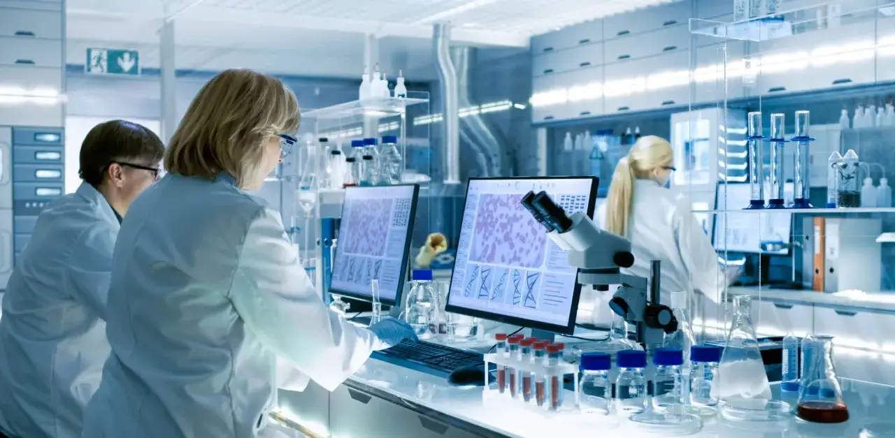 Naukowcy w białych fartuchach analizują dane na ekranach, co bada biolog. Mikroskop i probówki sugerują badania komórkowe.