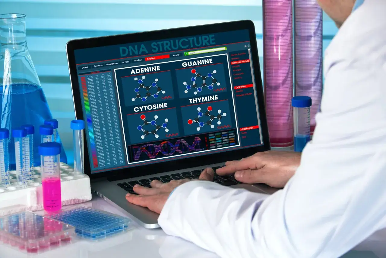 Biolog bada strukturę DNA na ekranie laptopa, otoczony probówkami z kolorowymi płynami.