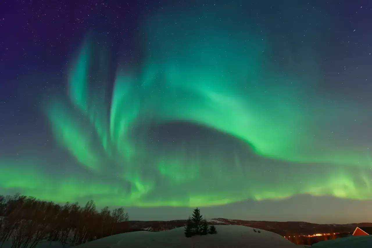 Zielona zorza polarna tańczy na nocnym niebie nad ośnieżonym krajobrazem. To widok, który sprawia, że zastanawiasz się, ile trwa zima w Norwegii.