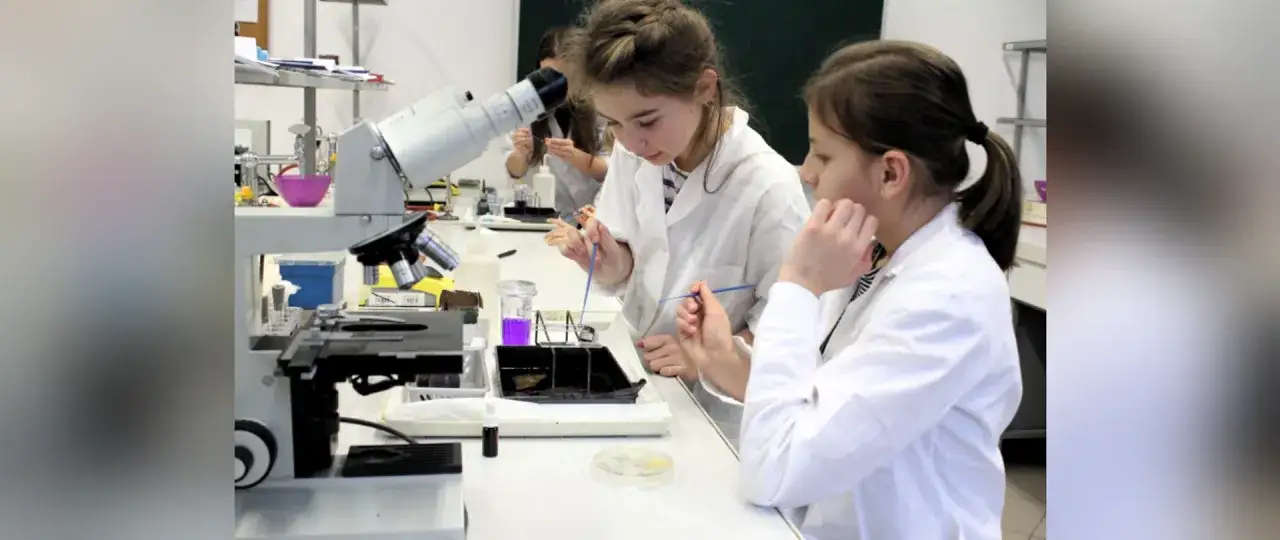 Dwie dziewczynki w kitlach laboratoryjnych badają pod mikroskopem próbki. To fascynujące, co bada biolog!