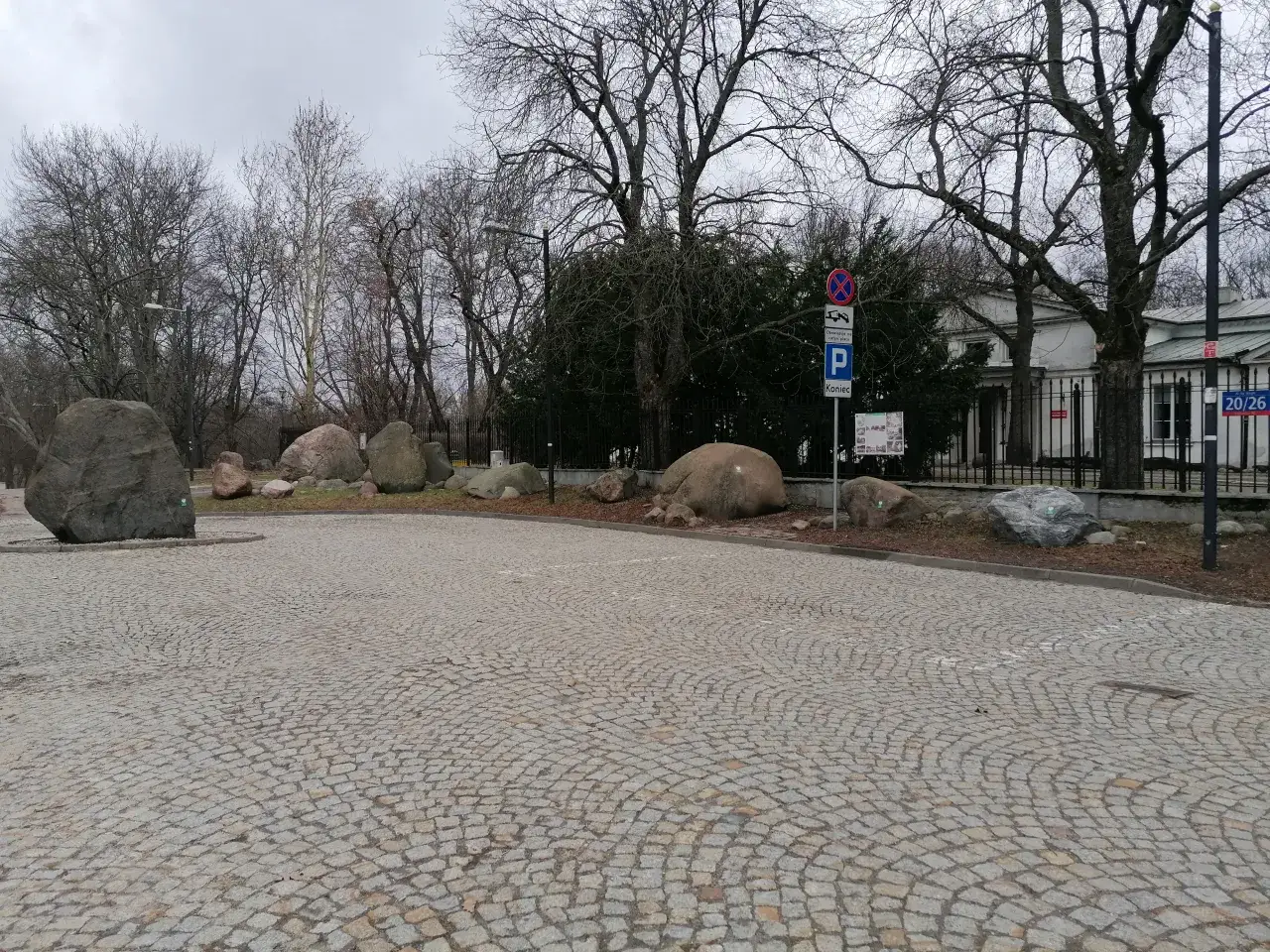 Kamienie polodowcowe na brukowanym placu, jakby pozostałości ostatniej epoki lodowcowej. Drzewa bez liści, szare niebo.